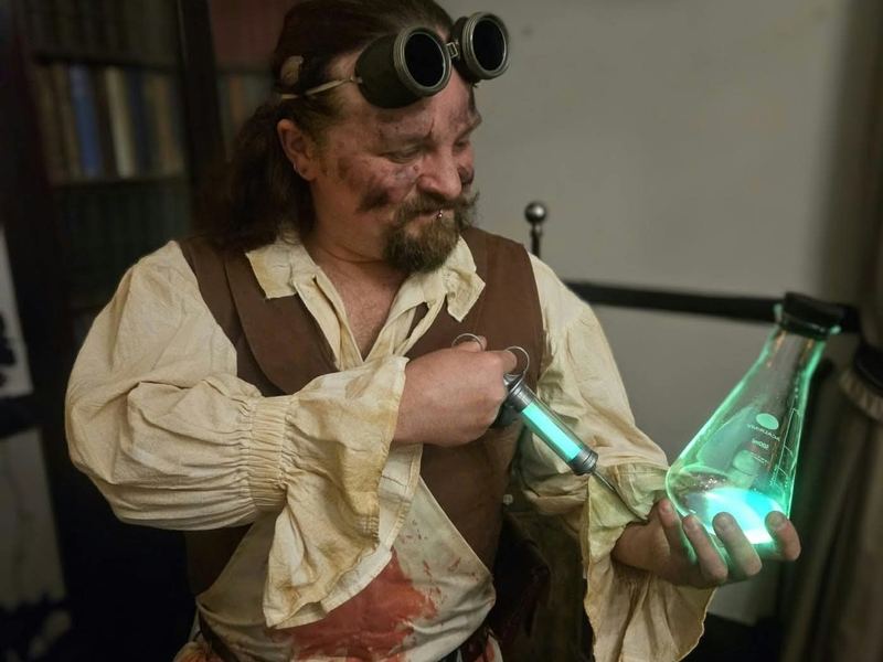 The Doctor in goggles and a blood-stained apron studying a syringe and a glowing green flask at the Reanimation Dinner