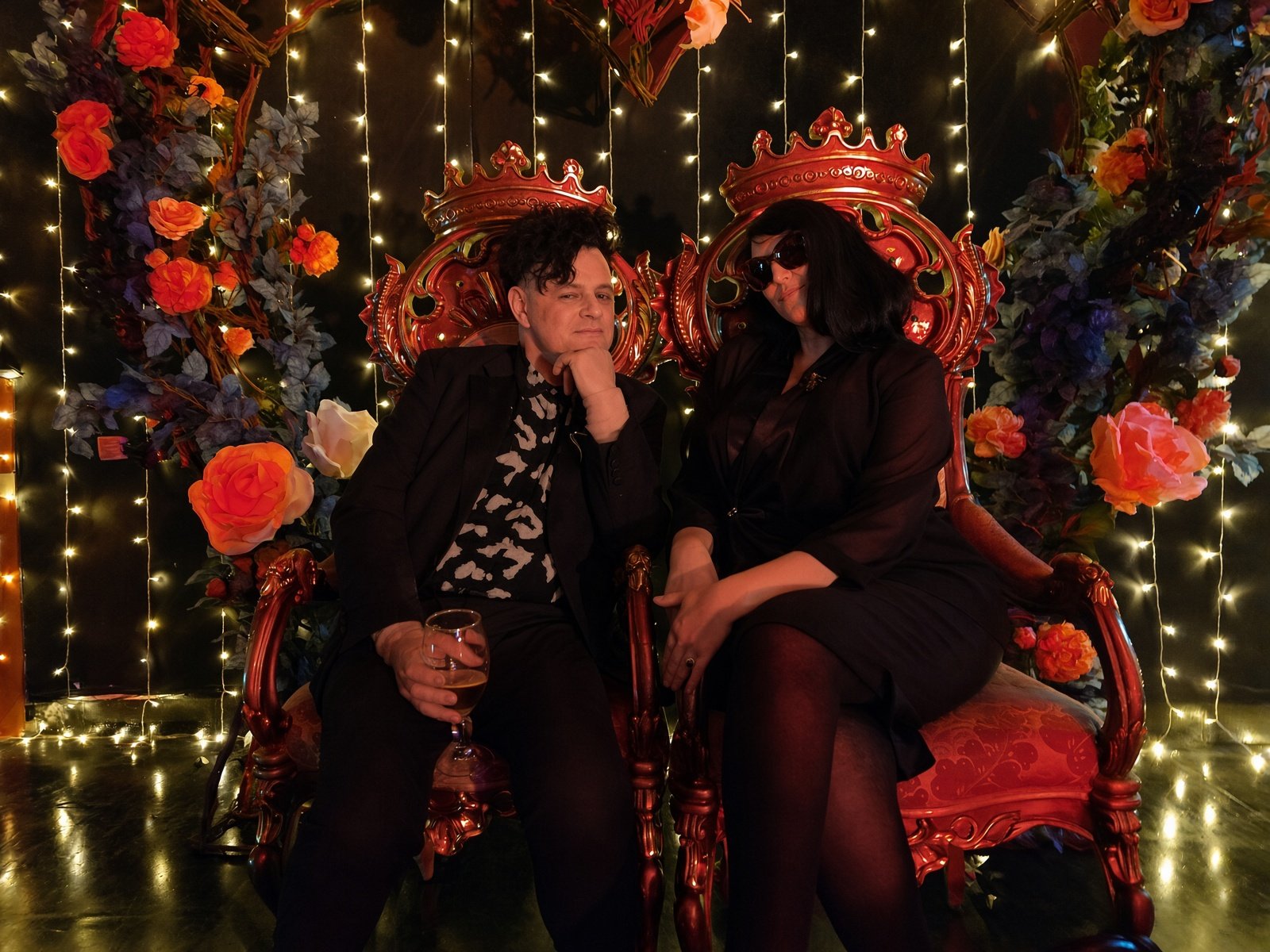 Two guests in black attire seated together on the ornate red and gold thrones at Underland, beneath a heart-shaped arch of roses and fairy lights
